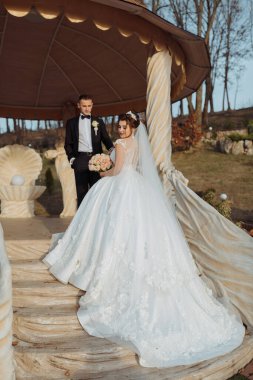 Gelin ve damat, beyaz elbiseli gelin ve siyah takım elbiseli damatla birlikte bir köşkün önünde duruyorlar.