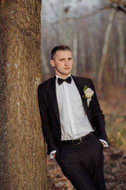 Siyah takım elbiseli ve beyaz gömlekli bir adam bir ağacın yanında duruyor. Siyah bir papyon ve yaka çiçeği takıyor. Adam bir fotoğraf için poz veriyor, ve ormanlık bir alan.