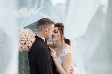 Bir gelin ve damat beyaz bir perdenin önünde öpüşüyor. Gelin beyaz bir elbise giyiyor ve damat siyah bir takım giyiyor.