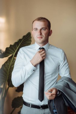 Beyaz gömlekli ve siyah kravatlı bir adam bir bitkinin önünde duruyor. Kravatını düzeltiyor ve bir ceket tutuyor. Formalite ve profesyonellik kavramı. Çünkü adam takım elbise ve kravat giymiş.