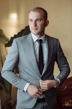 Gri takım elbiseli ve siyah kravatlı bir adam bir bitkinin önünde duruyor. Kravatını düzeltiyor ve kameraya bakıyor. Adam takım elbise giydiği için resmiyet ve profesyonellik kavramı.