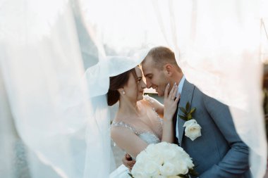 Bir gelin ve damat beyaz bir peçenin altında birbirlerini kucaklıyorlar. Gelin elinde bir buket beyaz çiçek tutuyor.