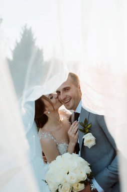 Bir gelin ve damat beyaz bir peçenin altında öpüşüyor. Gelin elinde bir buket beyaz çiçek tutuyor.