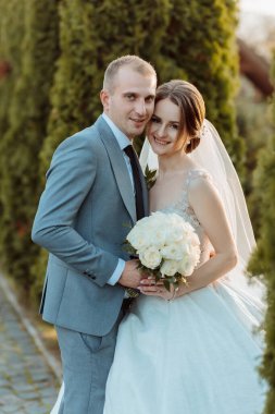 Bir gelin ve damat bir çalılığın önünde fotoğraf için poz veriyorlar. Gelin elinde bir buket beyaz çiçek tutuyor.