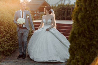 Bir gelin ve damat yolda yürüyorlar, gelin uzun beyaz bir elbise giyiyor. Adamın elinde bir buket çiçek var.