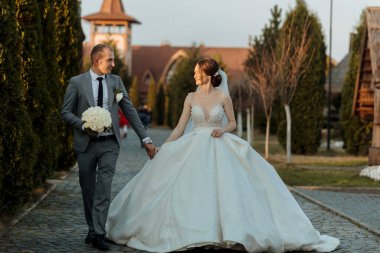 Bir gelin ve damat kaldırım taşı caddesinde yürüyor, el ele tutuşuyor ve gülümsüyor. Gelin uzun beyaz bir elbise giyiyor ve damat bir takım elbise giyiyor. Sahne romantik ve mutlu.