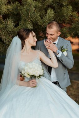 Bir gelin ve damat bir ağacın önünde fotoğraf için poz veriyorlar. Gelin bir buket tutuyor ve damat elini tutuyor. Sahne mutlu ve romantik.