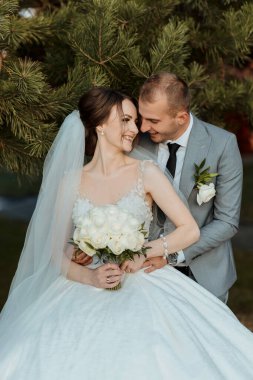 Bir gelin ve damat bir ağacın önünde fotoğraf için poz veriyorlar. Gelin beyaz bir elbise giyiyor ve elinde bir buket çiçek tutuyor. Damat takım elbise ve kravat takıyor. Sahne mutlu ve romantik.