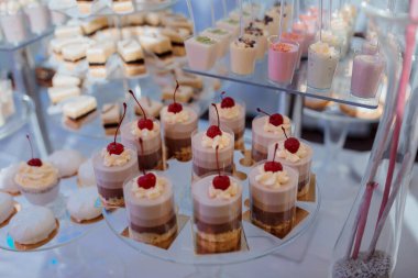 Pasta ve kekler de dahil olmak üzere çeşitli tatlılar içeren bir tatlı masası. Tatlılar cam bir tabakta sıralanır ve kirazlarla kaplanır. Masa bir parti ya da etkinlik için hazırlanmıştır.