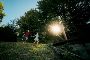 Bir çift ormanda el ele tutuşarak koşuyor. Manzara neşeli ve eğlenceli.
