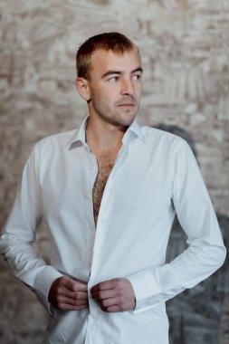 Beyaz gömlekli bir adam bir duvarın önünde duruyor. Düğmeleri açık bir gömlek giyiyor ve gömleğinin yakası açık.