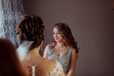 İki kadın aynanın önünde dikiliyorlar, biri gelinlik giyiyor. Diğer kadın yansımasına bakıyor ve gülümsüyor. Sahne mutlu ve kutlama dolu.