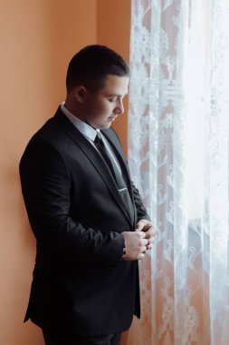 Takım elbiseli bir adam pencerenin önünde duruyor, kravatını düzeltiyor. Formalite ve profesyonellik kavramı. Çünkü adam takım elbise ve kravat giymiş.