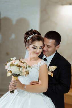 Bir gelin ve damat bir buket çiçek ve yaka çiçeği tutarken birbirlerini kucaklıyorlar. Gelin beyaz bir elbise giyiyor ve damat siyah bir takım elbise giyiyor. Sahne romantik ve mutlu.