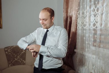 Beyaz gömlekli ve siyah kravatlı bir adam saatine bakıyor. O bir kravat ve beyaz bir gömlek giyiyor.