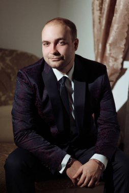 Takım elbiseli ve kravatlı bir adam kanepede oturur. Siyah bir ceket ve beyaz bir gömlek giyiyor.