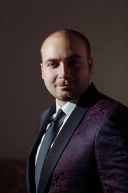 Takım elbiseli ve kravatlı bir adam kameraya gülümsüyor. Adam mor ve pembe desenli siyah bir takım elbise giyiyor.