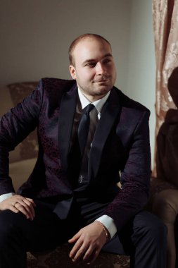 Takım elbiseli ve kravatlı bir adam kanepede oturuyor. Gülümsüyor ve kameraya bakıyor.