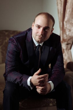 Takım elbiseli ve kravatlı bir adam kanepede oturur. Mor bir ceket ve beyaz bir gömlek giyiyor.