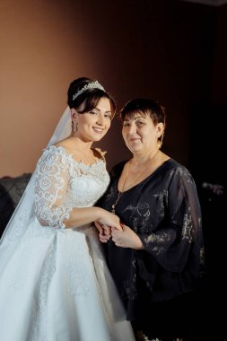Bir gelin ve annesi birlikte duruyorlar, gelin beyaz bir elbise giyiyor ve anne siyah bir elbise giyiyor. El ele tutuşuyorlar, muhtemelen birbirlerine sevgilerini ve desteklerini göstermek için.