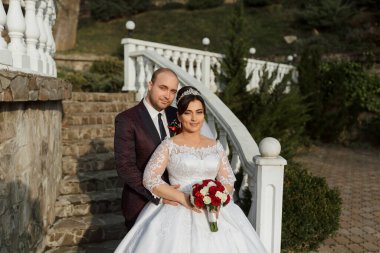 Bir gelin ve damat merdivende fotoğraf çektirmek için poz veriyorlar. Gelin beyaz bir elbise giyiyor ve damat bir takım elbise giyiyor. Gelin elinde bir buket çiçek tutuyor.