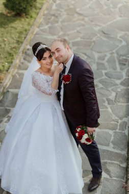 Bir gelin ve damat taş bir merdivende fotoğraf çektirmek için poz veriyorlar. Gelin beyaz bir elbise giyiyor ve damat siyah bir takım elbise giyiyor. Gelin damadın boynuna dokunuyor.