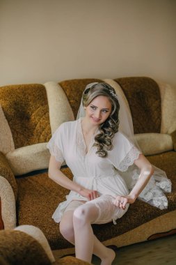 Beyaz elbiseli bir kadın kanepeye oturmuş, duvak takmış ve beyaz bir duvak takmış. Gülümsüyor ve düğüne hazırlanıyor.