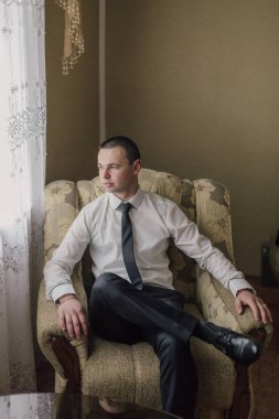 Takım elbiseli ve kravatlı bir adam sandalyede oturur. O, pencereden dışarı bakıyor. Sandalye kahverengi ve çiçek desenli.