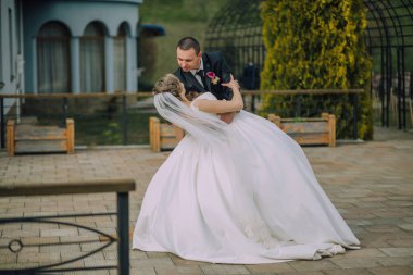 Bir gelin ve damat tuğla bir verandada birbirlerini kucaklıyorlar. Gelin beyaz bir elbise giyiyor ve damat bir takım elbise giyiyor. Sahne mutlu ve romantik.