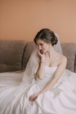 Beyaz elbiseli bir kadın kanepede elleri yüzünde oturuyor. Duvak ve kolye takıyor.