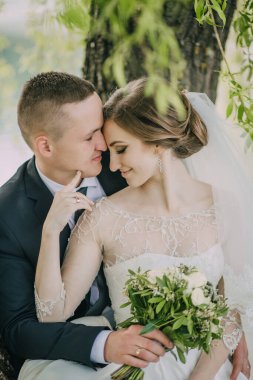 Bir gelin ve damat bir ağacın altında oturuyor, el ele tutuşuyor ve birbirlerinin gözlerine bakıyor.