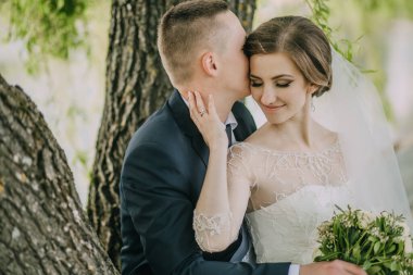 Bir gelin ve damat bir ağacın altında oturmuş, birbirlerini öpüyorlar. Gelin elinde bir buket çiçek tutuyor.