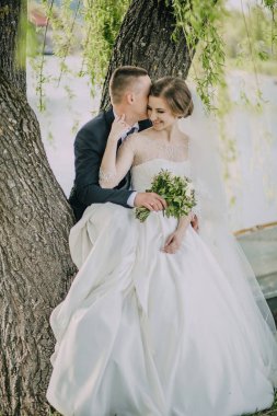 Gelin ve damat bir ağacın altında oturuyor, gelin bir buket tutuyor. Sahne romantik ve samimi.