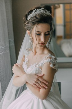 Gelinlik giymiş bir kadın taç takıyor ve kolunu tutuyor. Kameraya bakıyor.