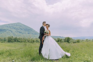 Bir gelin ve damat, arka planda güzel bir dağ manzarası olan bir tarlada duruyorlar.