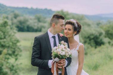 Gelin ve damat bir tarlada, gelin bir buket çiçek tutuyor. Sahne romantik ve mutlu.