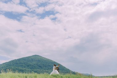 Bir gelin ve damat arka planda bir dağ ile, çimenli bir yamaçta duruyorlar. Gökyüzü bulutlu, ama çift anın tadını çıkarıyor gibi görünüyor.
