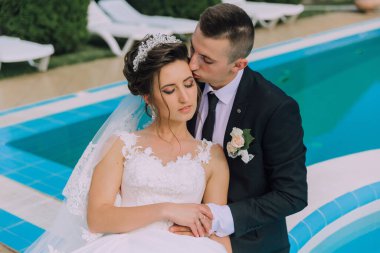 Gelin ve damat havuzda oturuyor, gelin uzanıyor ve damat onu öpüyor.
