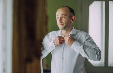 Bir adam pencerenin önünde giyiniyor. Beyaz bir gömlek ve kravat takıyor.