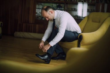 Bir adam sarı bir sandalyede oturuyor ve ayakkabılarını tamir ediyor. Beyaz gömlek ve mavi pantolon giyiyor.