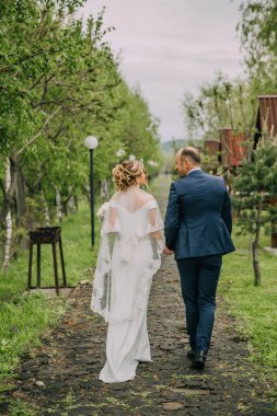 Bir gelin ve damat ormanda bir patikada yürüyorlar. Gelin beyaz bir elbise giyiyor ve damat mavi bir takım giyiyor.