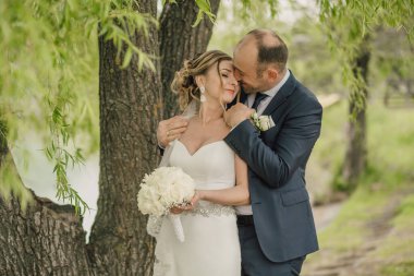 Bir adam ve bir kadın bir ağacın altında sarılıyorlar. Adam mavi bir takım giyiyor ve kadın beyaz bir elbise giyiyor.