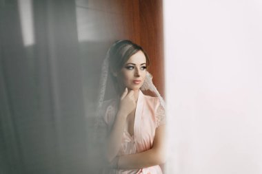 Pembe bornozlu bir kadın pencereden dışarı bakıyor. Peçe takıyor ve elleri kalçasında.