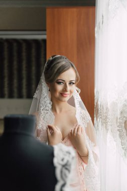 Gelinlik giymiş bir kadın mankenin önünde dikiliyor. Peçe takıyor ve gülümsüyor.