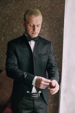 Siyah takım elbiseli bir adam resmi bir etkinliğe hazırlanıyor. Kravatını düzeltiyor ve saatini kontrol ediyor. Özel bir durum için formalite ve hazırlık kavramı