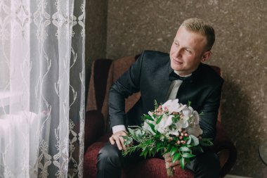 Takım elbise giyen ve kravat takan genç bir adam kucağında bir buket çiçekle bir sandalyeye oturur. Çiçekler beyaz ve taze gibi görünüyor. Adam çiçeklerden hoşlanıyor ve gülümsüyor.