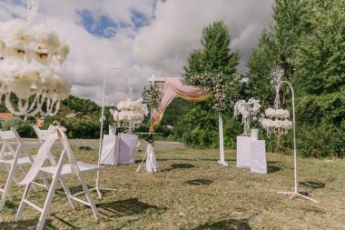 Beyaz sandalyeli ve beyaz kemerli bir tarlada bir düğün töreni yapılacak. Sandalyeler kemerin etrafında yarım daire şeklinde dizilmiş ve birkaç vazo var. Atmosfer çok zarif.