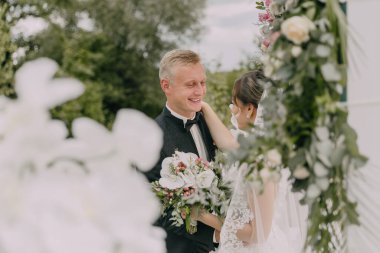 Bir gelin ve damat beyaz bir kemerin önünde duruyor. Gelin bir buket çiçek tutuyor ve damat bir buket çiçek tutuyor. Birbirlerine gülümsüyorlar.