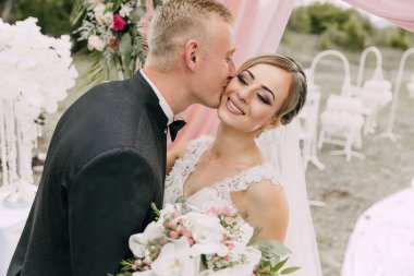 Gelin ve damat beyaz bir çadırın önünde öpüşürler. Gelin beyaz bir elbise giyiyor ve damat siyah bir takım elbise giyiyor. Sahne bir bahçede geçiyor, yakınlarda sandalyeler ve masa var.