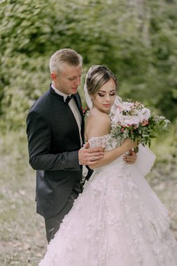 Bir gelin ve damat ormanda fotoğraf çektirmek için poz veriyorlar. Gelin elinde bir buket çiçek tutuyor.